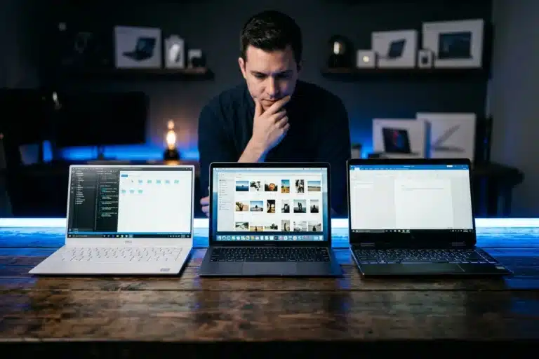 Pessoa comparando notebooks em 2026 para escolher o modelo certo: vários notebooks abertos sobre uma mesa com iluminação azul