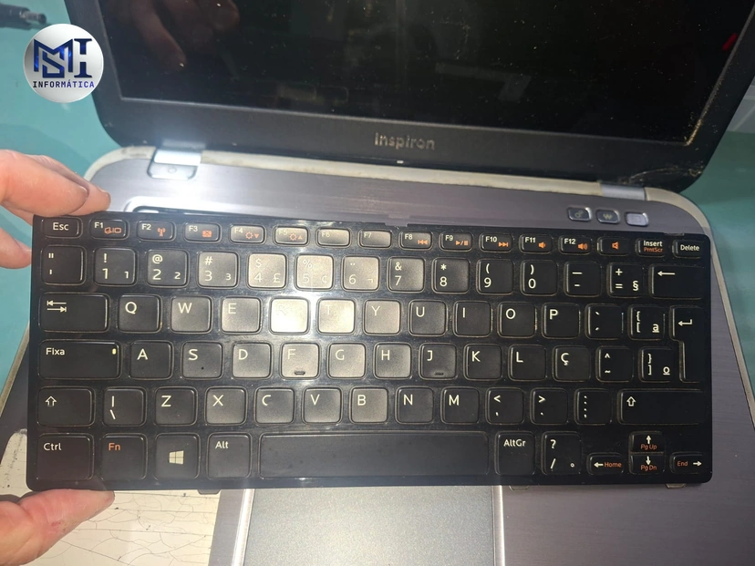 Teclado ABNT de um notebook Dell Inspiron retirado e segurado na mão, com o notebook aberto atrás