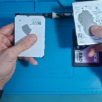 Dois HDs de notebook nas mãos do técnico e, embaixo, um SSD SATA de 2,5 polegadas