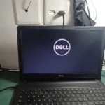 Notebook Dell aberto numa mesa com o logo da Dell na tela, ao lado do multímetro, no vídeo do canal