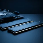 Dois pentes de memória RAM de desktop com dissipador escuro sobre uma mesa, ao lado de uma placa-mãe desfocada