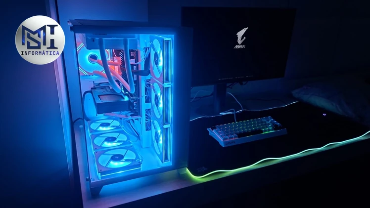 PC gamer de gabinete branco iluminado em azul ao lado de um monitor AORUS, com teclado mecânico compacto sobre um mousepad grande com luz na borda