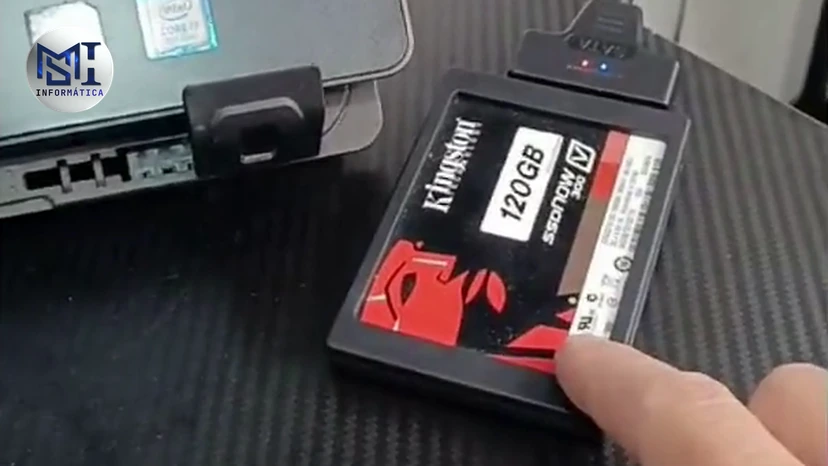Dedo do técnico apontando um SSD de 2,5 polegadas num adaptador USB ligado a um notebook, como um disco externo, no vídeo do canal